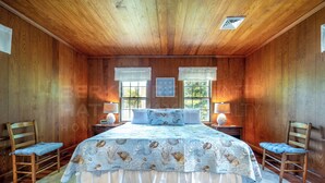 Une chambre avec un plafond en bois, deux fenêtres avec des rideaux, un lit recouvert d’une couvre-lit à motifs, deux tables de chevet avec des lampes et deux chaises.
