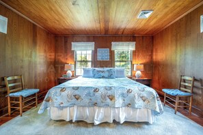 Une chambre avec un plafond en bois, deux fenĂȘtres avec des rideaux, un lit recouvert dâune couvre-lit Ă motifs, deux tables de chevet avec des lampes et deux chaises.