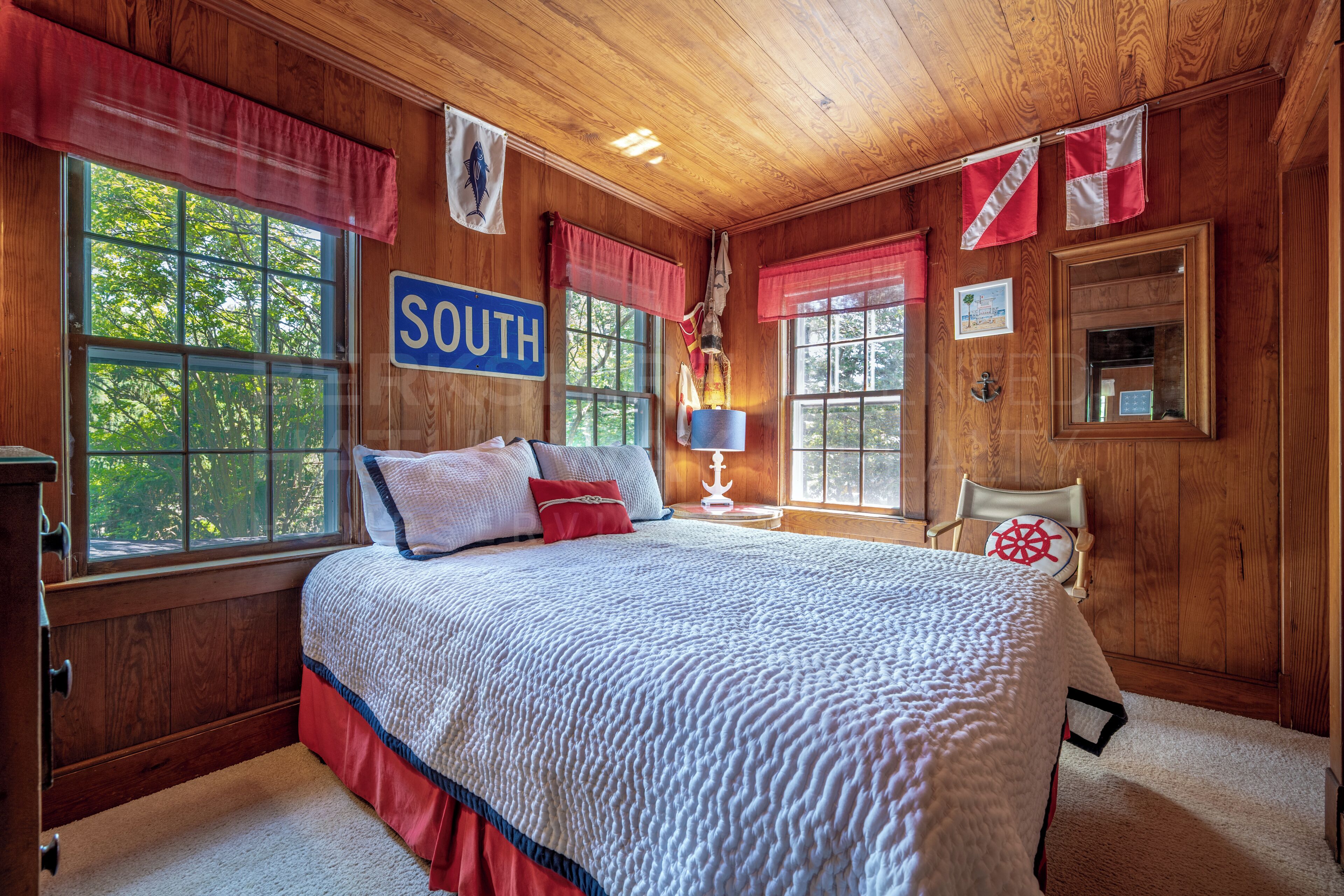 A wooden-paneled bedroom with a large bed, a nightstand, a chair, and a window with a view of trees.