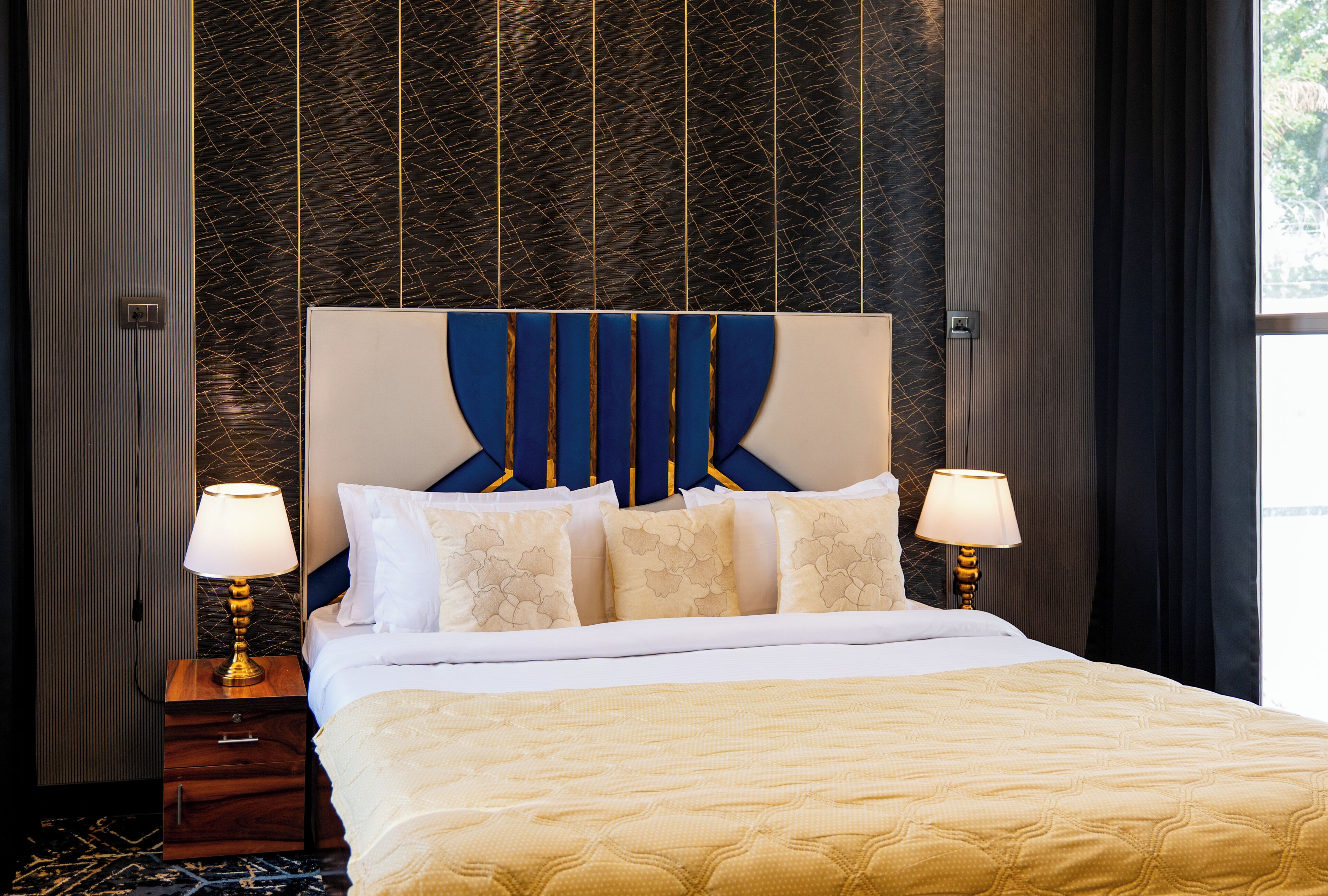 Una habitación de hotel con una cama, dos mesitas de noche con lámparas, un tocador de madera y un papel tapiz estampado.