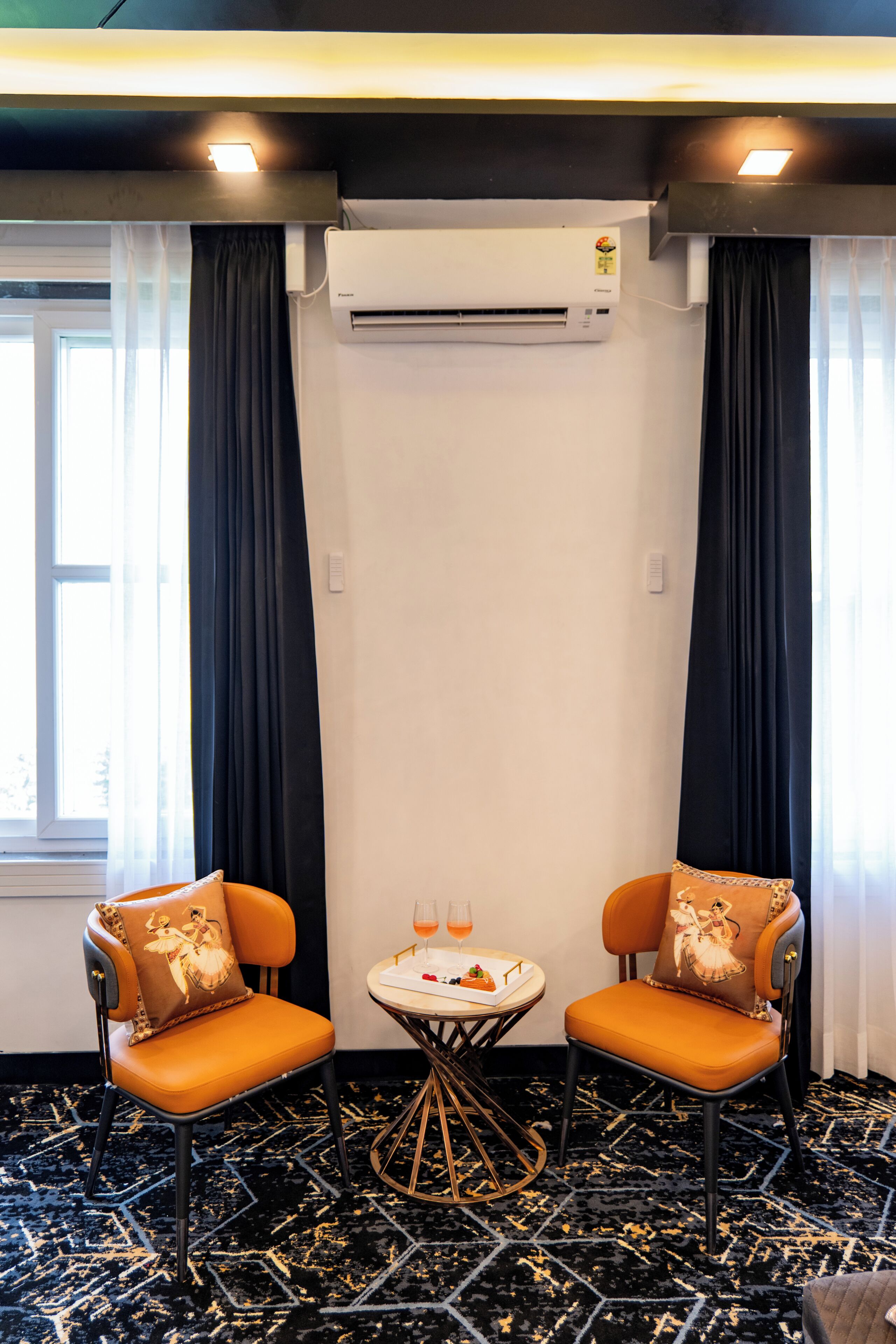 Habitación de hotel con dos sillas naranjas, una mesita redonda con un vaso de jugo de naranja, y un aire acondicionado en la pared.