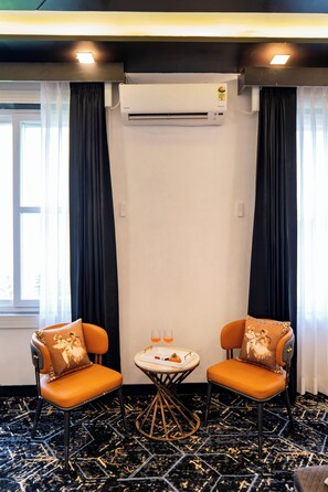 Et hotelværelse med to orange farvede stole, et rundt bord med et glas appelsindrik, og en aircondition på væggen.