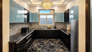 A modern kitchen with a black marble countertop, a central island, and a chandelier. - ELIVAAS Signature Raj Farms | 3 BHK Villa with Pvt Pool (Gurugram)