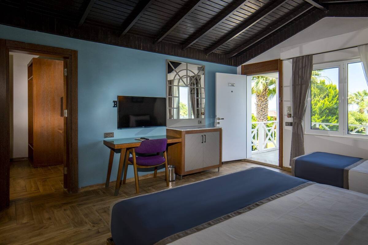 A room with a bed, a desk with a chair, a TV, a wardrobe, and a balcony with a view of greenery.