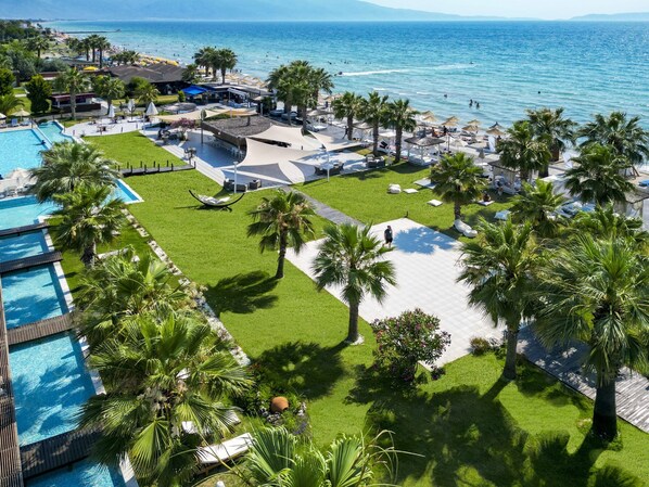A resort with a swimming pool, lounge areas, palm trees, and a view of the sea.