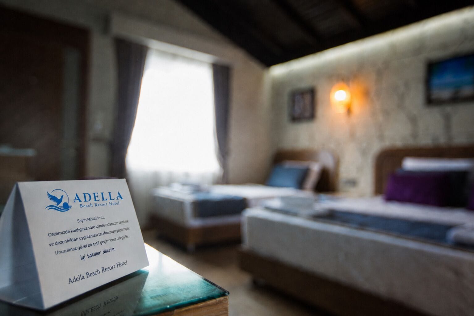 A hotel room with two beds, a window, and a sign for Adella Beach Resort Hotel.