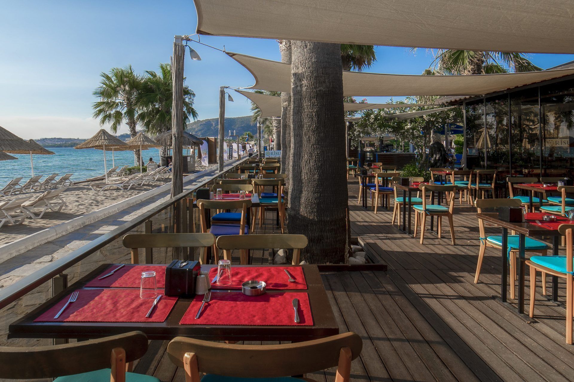 A beachside restaurant with outdoor seating, tables with red placemats, and a view of the sea.