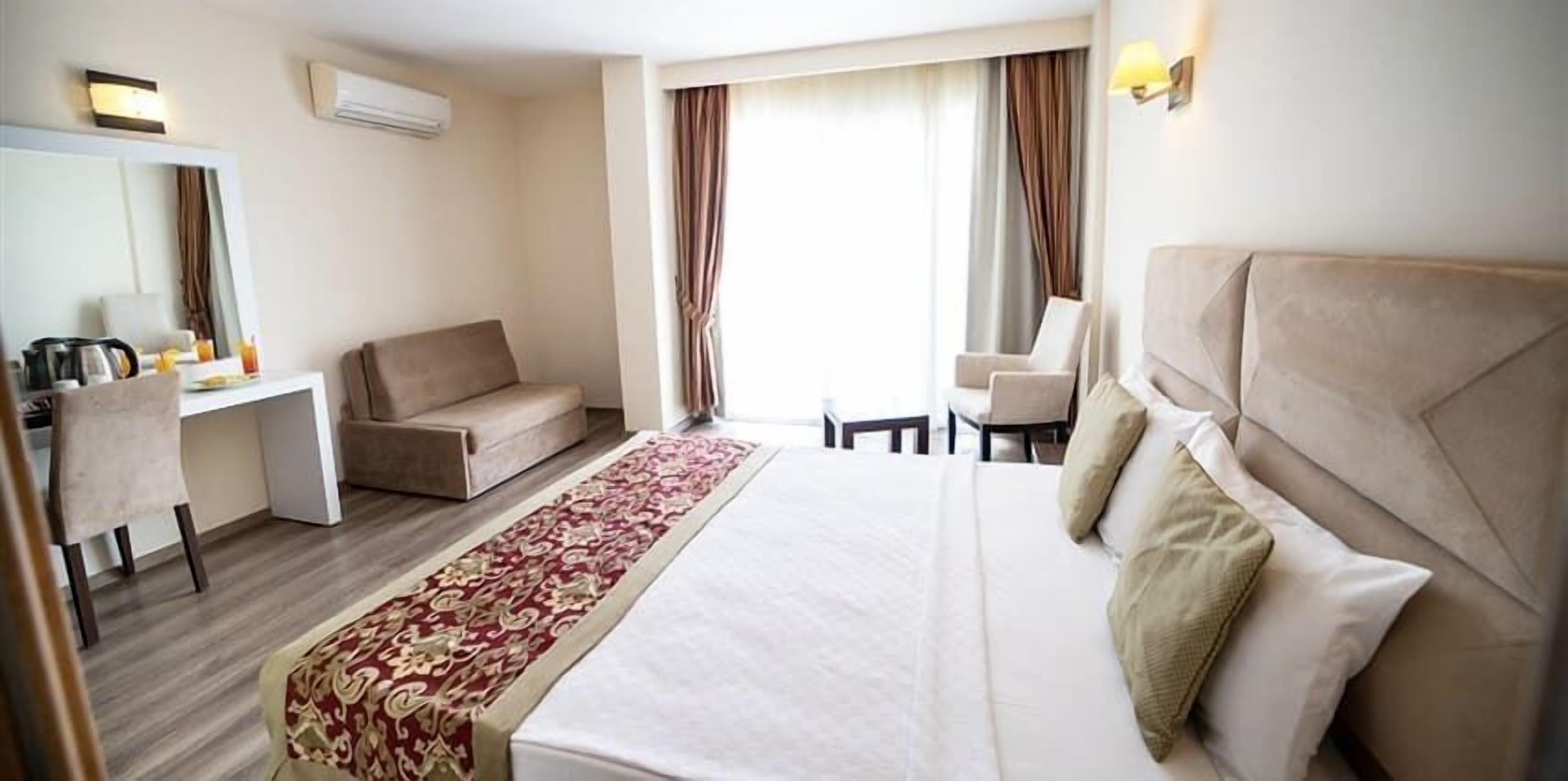 A hotel room with a bed, a desk, a chair, a sofa, a long red and white patterned runner, and a large window with curtains.