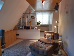 A cozy room with a brown leather sofa, a glass side table, a wooden shelf with decorative items, and a window with curtains.