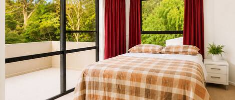 A bedroom with a large window, a bed with a plaid bedspread, red curtains, and a nightstand with a plant.
