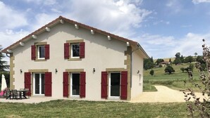 Une maison à deux étages, avec une façade de couleur beige, des volets rouges et une terrasse aménagée avec du mobilier d’extérieur.