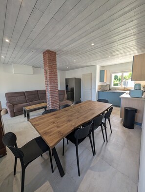 Een moderne keuken met een inbouwoven, een eethoek met een houten tafel en stoelen, en een zithoek met een bank en een bakstenen pilaar.