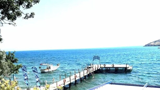 A pier extending into calm blue waters, a boat docked at the end, and a building with a blue roof in the foreground. - ÇÖKERTME BUTİK OTEL (Milas)