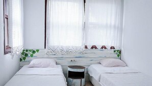 Quarto com duas camas, uma mesa pequena, uma janela com cortinas e uma planta.