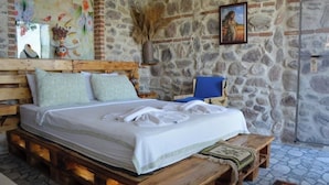 Uma cama com colchão e almofadas brancas, cabeceira de madeira e um banquinho ao lado.