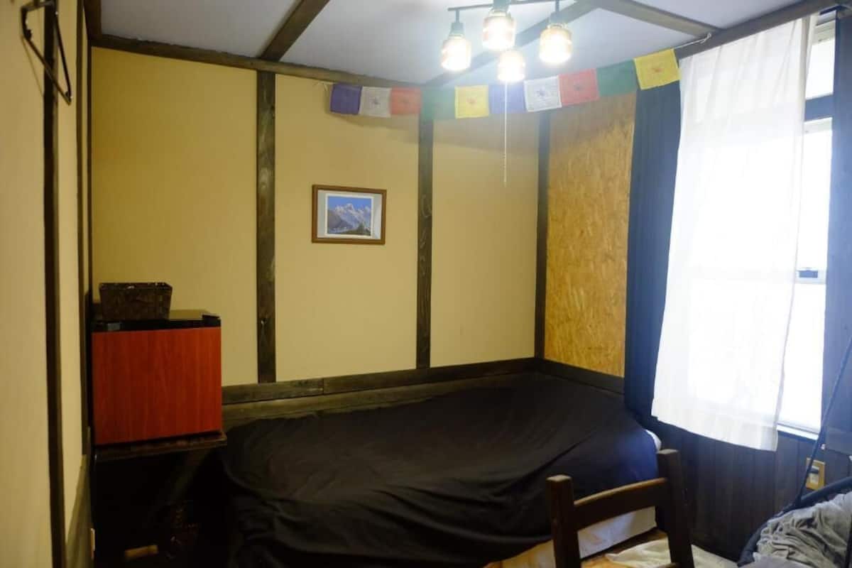 A room with a bed, a small table with a box, a framed picture on the wall, and a window with curtains.