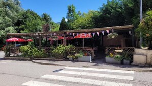 A covered outdoor seating area with tables and umbrellas, surrounded by potted plants and trees. - L'étape (Les Omergues)