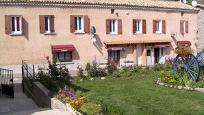 A hotel with a tiled roof, multiple windows with shutters, a garden with flowers and a wheelbarrow, and a sign indicating a restaurant. - L'étape (Les Omergues)