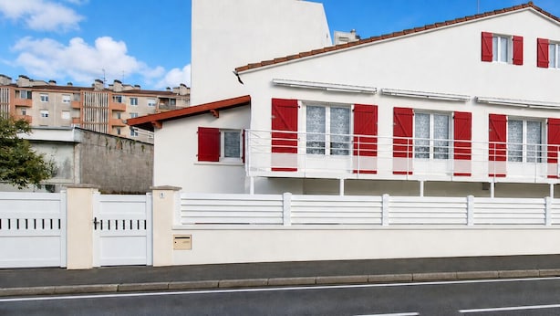 A two-story building with red shutters, a white facade, and a gated entrance.