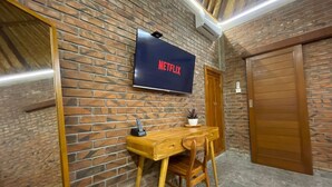 Une pièce avec un mur en briques, un bureau en bois, un téléviseur affichant le logo « NETFLIX », et une porte en bois.