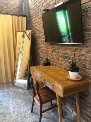 Een kamer met een bakstenen muur, een houten bureau, een stoel, een aan de muur bevestigde televisie en een potplant op het bureau.
