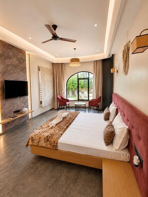 A modern hotel room with a large bed, a ceiling fan, a TV, a fireplace, a dining area with chairs, and a view of greenery outside. - Nakshatra Aura (Indore)