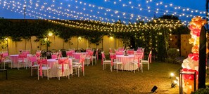 Un espacio para eventos al aire libre con mesas y sillas dispuestas para los invitados, luces colgantes y un arco decorativo.