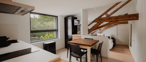 A modern kitchen with a large window, a dining area with a wooden table, and a staircase with exposed beams.