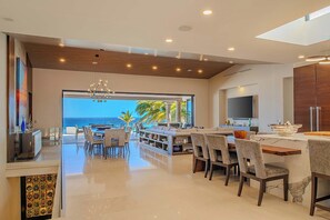 A modern kitchen with a large island, a dining area with chairs, and a view of the ocean. - Casa Keller: Beachfront in Pedregal | 2 Swimming Pools (Cabo San Lucas)