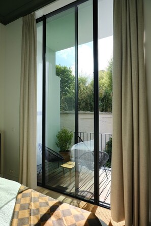 Ein Zimmer mit einem Bett, einem fensterbehangenen Fenster, einem Blick auf einen Balkon mit Stühlen und einer Pflanze sowie einer Glastür, die nach draußen führt.