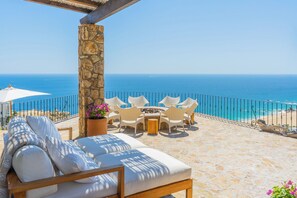Eine Steinterrasse mit einem weißen Sofa und Sesseln, einem kleinen Tisch und Blick auf das Meer.