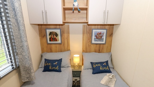 A small room with two single beds, each with a pillow that reads 'Bee kind.' There are framed pictures above the beds, a small shelf with a toy, and a window with blinds. - Plot 41 (Carlisle)