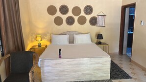 A hotel room with a bed, two bedside tables, a chair, and decorative wall hangings. - Heidi’s Beach Stay (Mombasa)