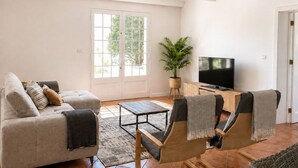 En stue med sofa, lænestole, sofabord, et fjernsyn på et træstel og et stort vindue med udsigt til grønne omgivelser.