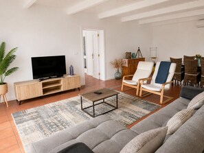 Een woonkamer met een bank, fauteuils, een salontafel, een tv op een houten standaard, en een eethoek met rieten stoelen.
