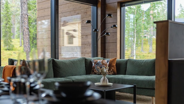 Un interior moderno con un sofá verde, una mesita con un florero y vistas a un bosque a través de grandes ventanales.