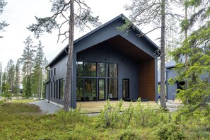 Una cabaña moderna con ventanas amplias, una terraza de madera y un bosque circundante.
