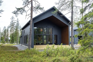 Una cabaña moderna con ventanas amplias, una terraza de madera y un bosque circundante.