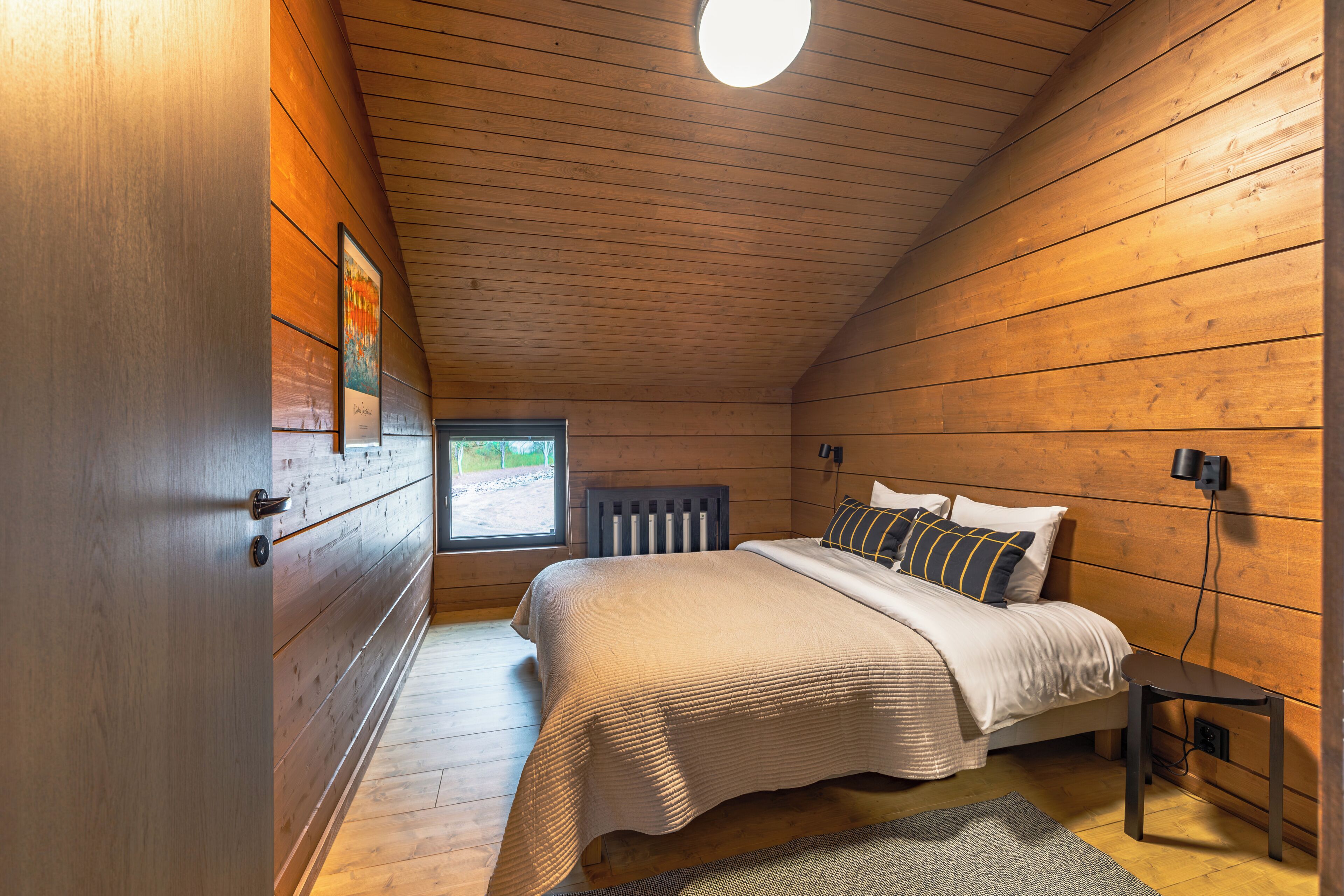 Een knus slaapkamer met een houten plafond, een bed met een beige deken, een nachtkastje en een raam met uitzicht op groen.
