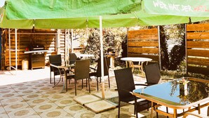 Une terrasse extérieure avec un parasol vert, des chaises en osier et une table avec un plateau en verre.
