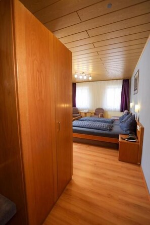 Quarto com guarda-roupa de madeira, cama com colchão azul, mesa de cabeceira e janela com cortinas.
