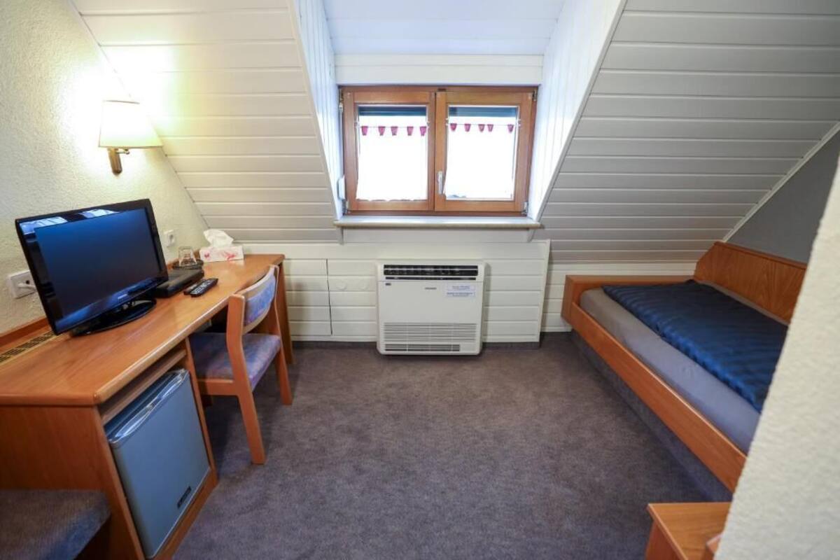 A compact room with a desk, a computer, a small refrigerator, a bed, and a window with curtains.