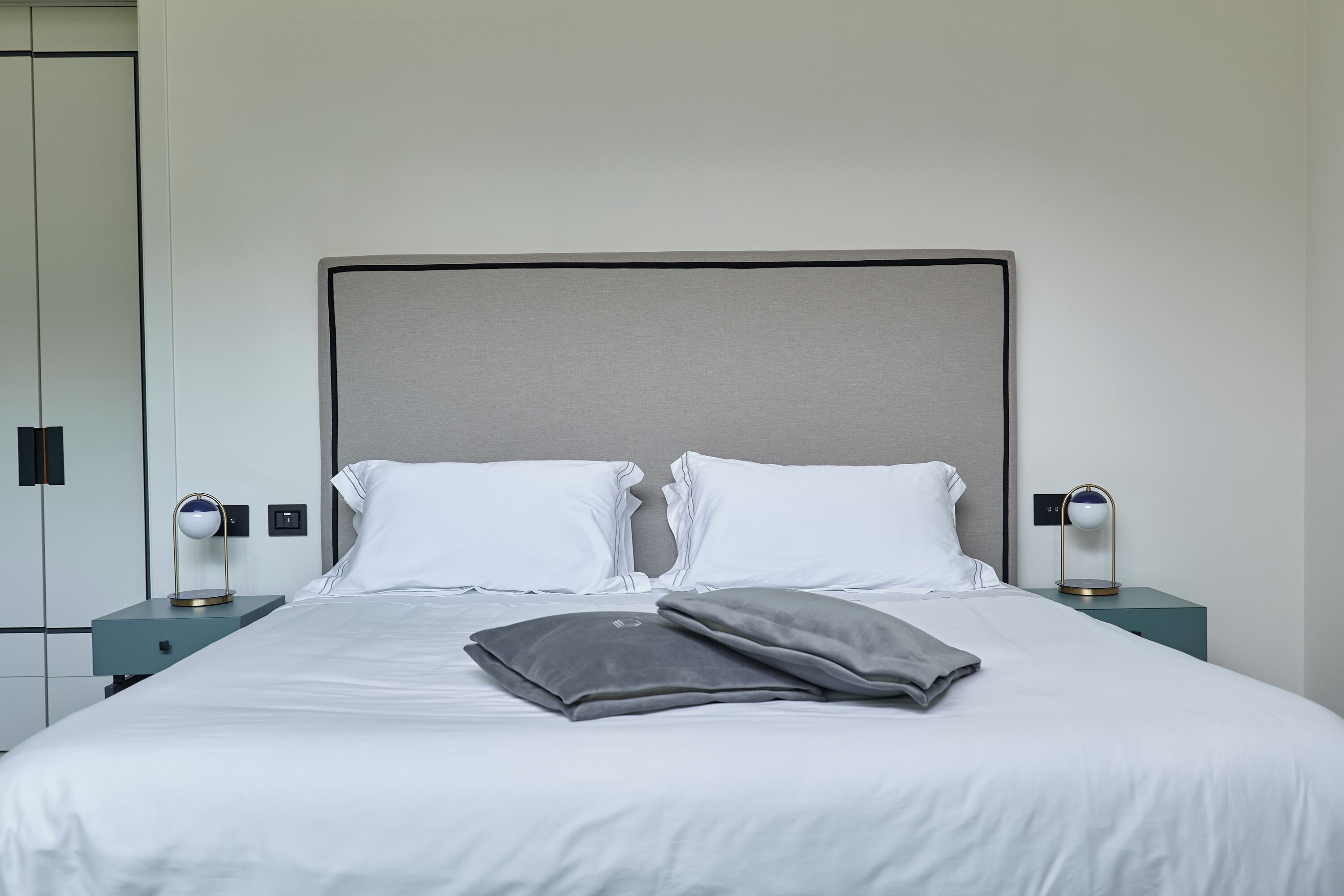 A neatly made bed with two pillows, a bedside table with a lamp, and a wall-mounted switch.
