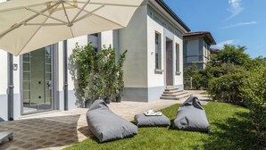 Un edifici amb façana blanca, un para-sol gran i seients exteriors amb coixins.