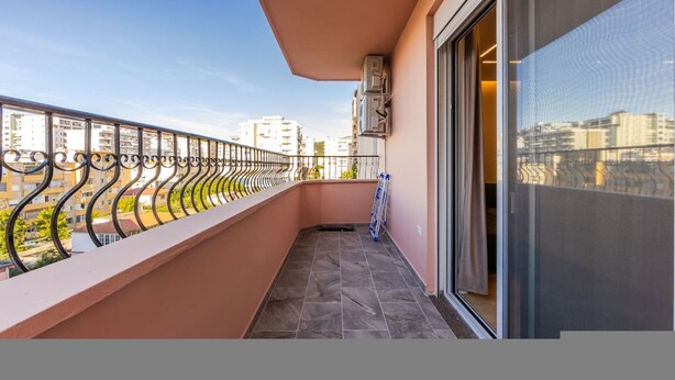 A balcony with a metal railing, a glass door, and a view of buildings. - A H Azure Apartaments 1BR (Vlorë)