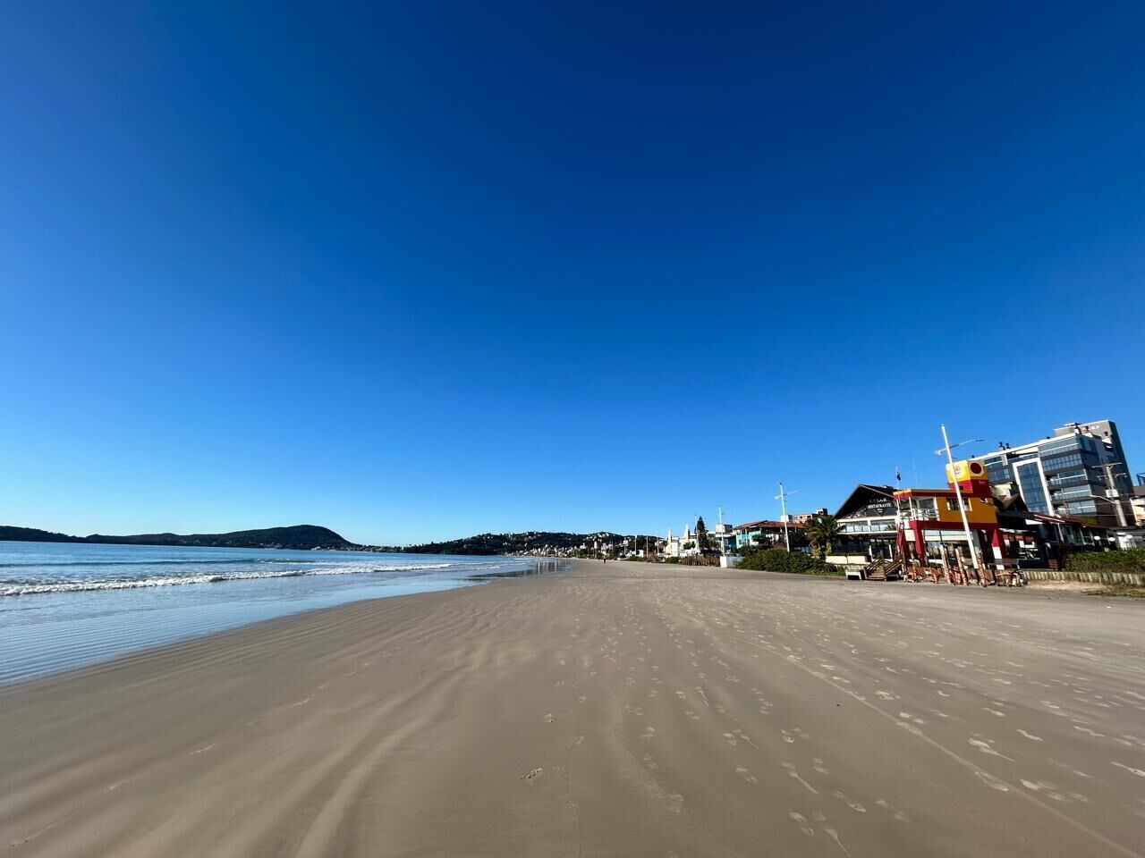 A beach with a clear sky, a sandy shore, and buildings in the background.