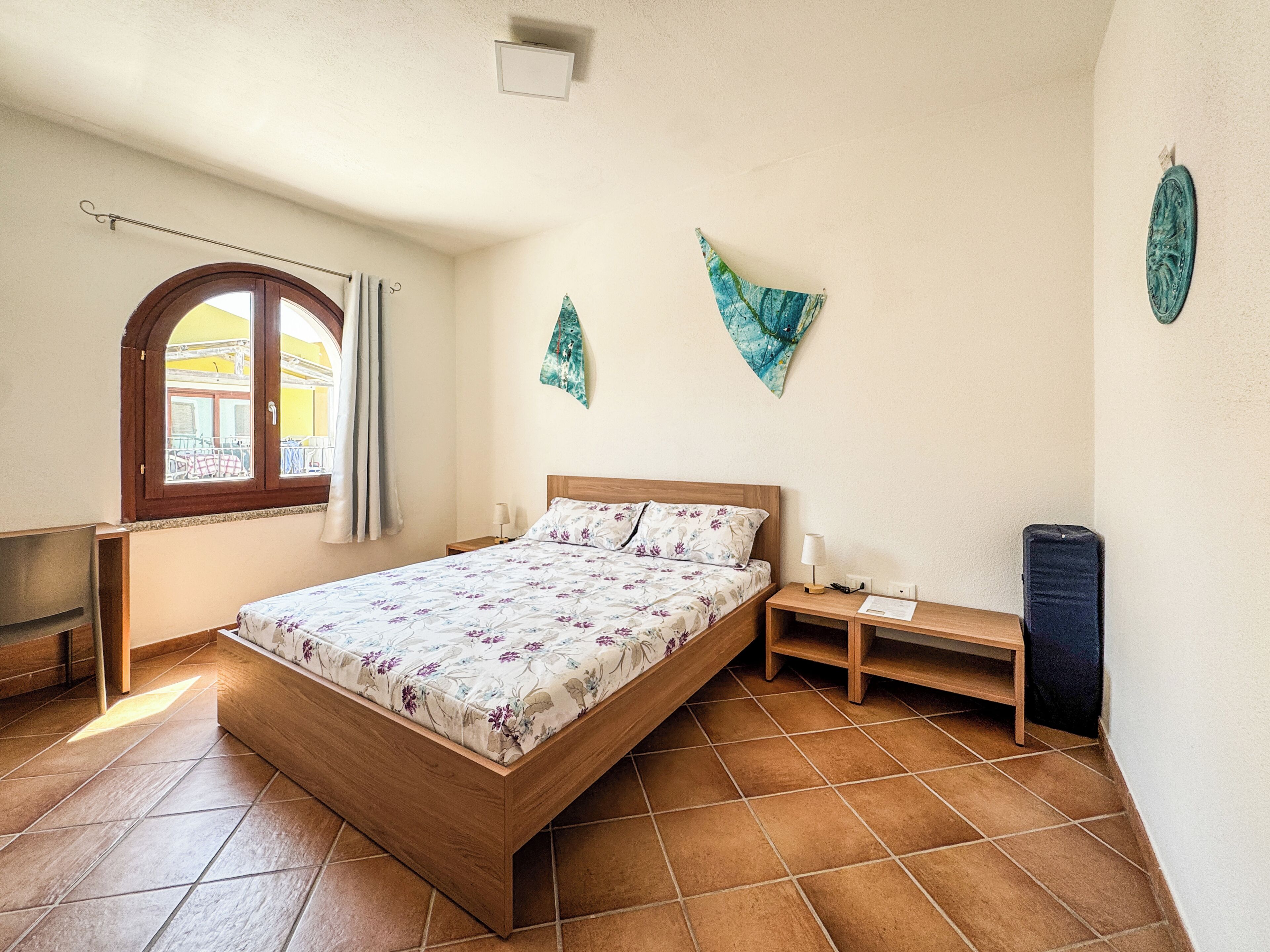 A bedroom with a wooden bed, a nightstand, a lamp, a window with curtains, and a wall-mounted decoration.