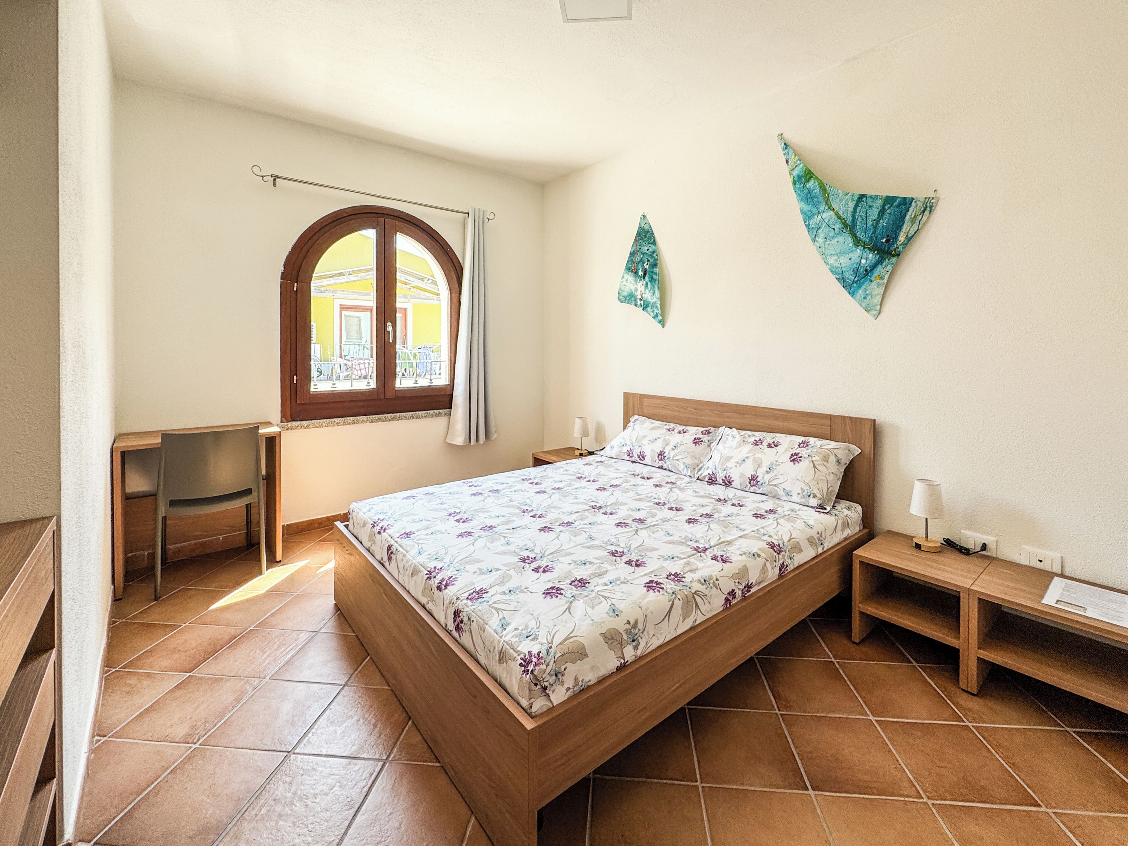 Una camera da letto con un letto, una scrivania, una sedia, una finestra con tende e un quadro appeso al muro.