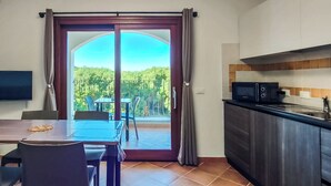 Ein Essbereich mit Tisch und Stühlen, einem Fernseher, einer Küche mit Mikrowelle und einer Glastür, die auf einen Balkon mit Blick ins Grüne führt.
