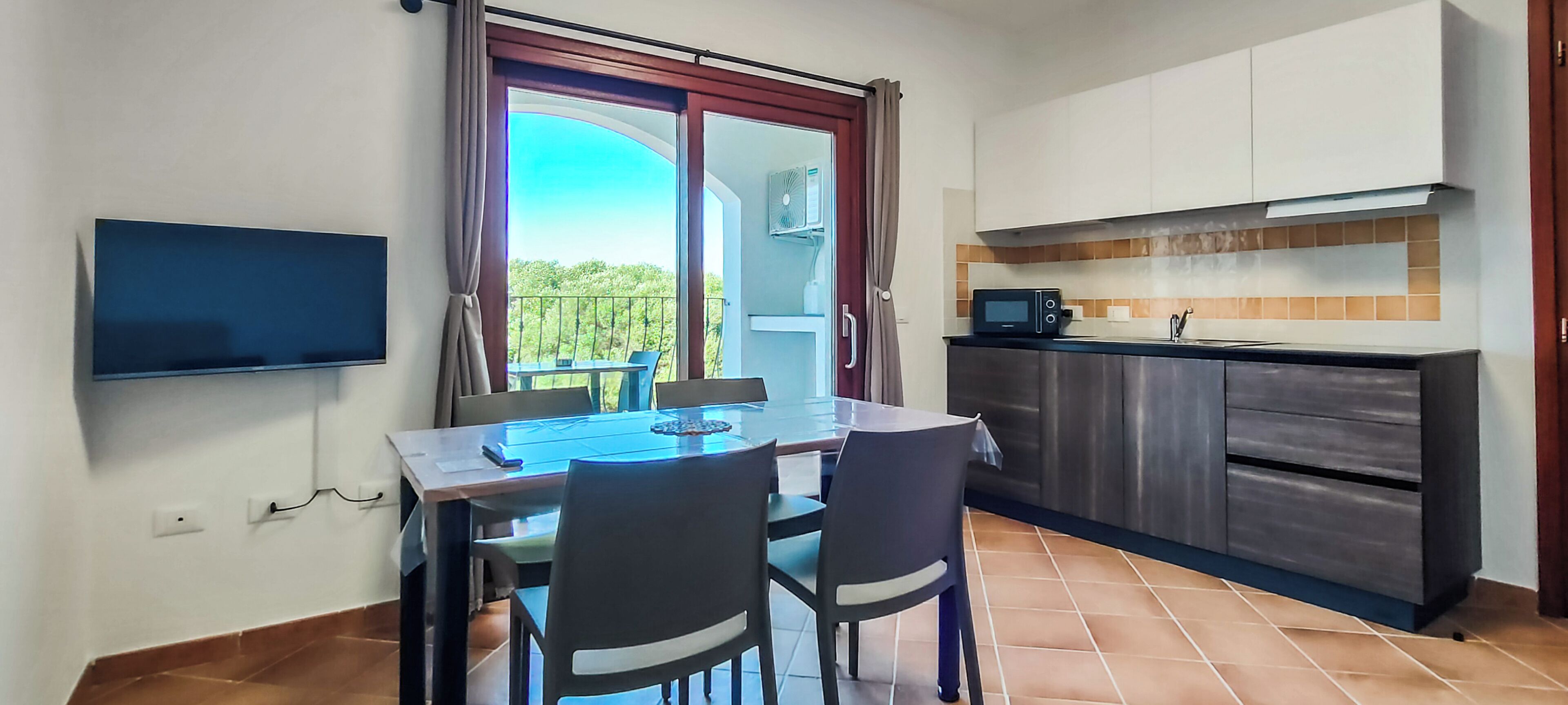 Un coin repas avec une table et des chaises, un téléviseur fixé au mur, une cuisine équipée d’un four à micro-ondes et une porte-fenêtre coulissante donnant sur un balcon.